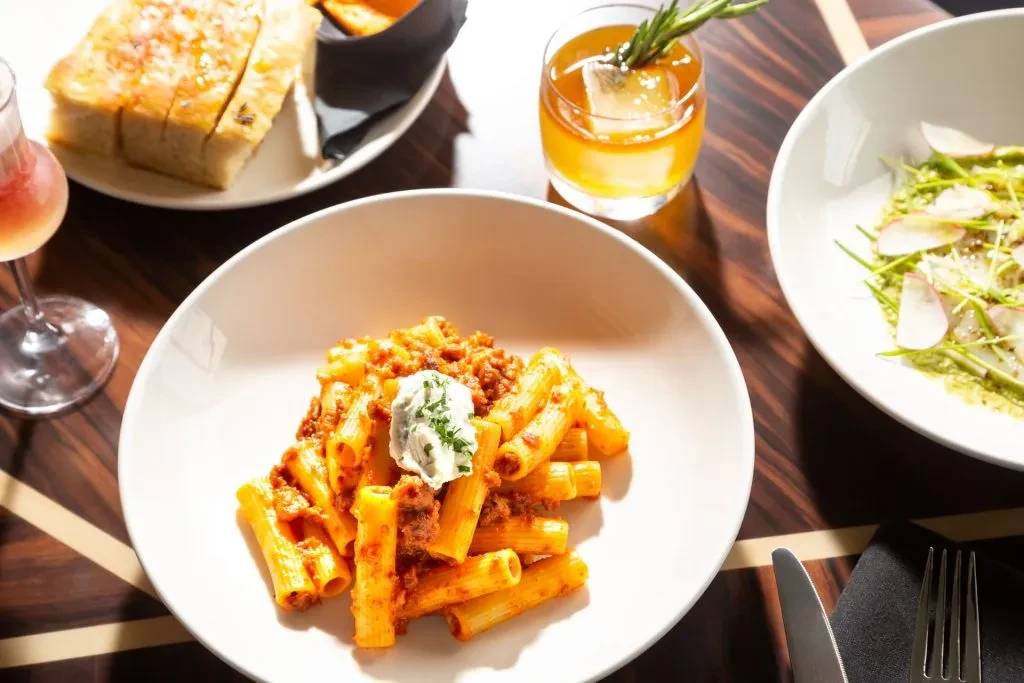 Rigatoni bolognese, focaccia, salad, and drinks on a dark wooden table.