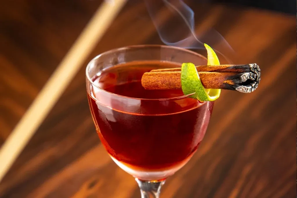 Reddish-brown cocktail with smoking cinnamon stick and green lime peel.