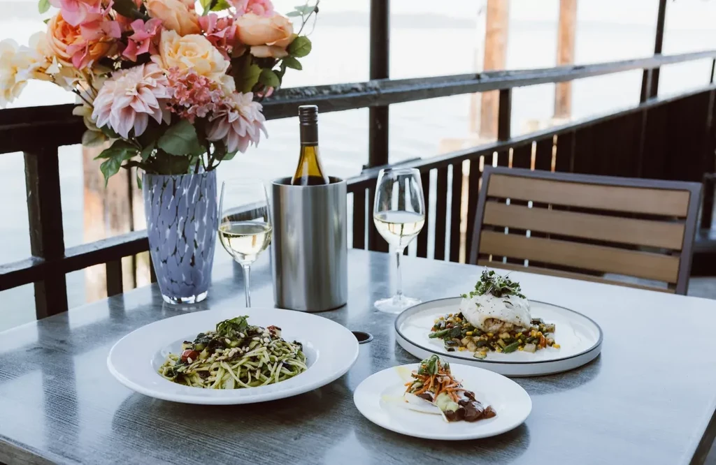 Outdoor dining table with food, white wine, flowers, overlooking water.