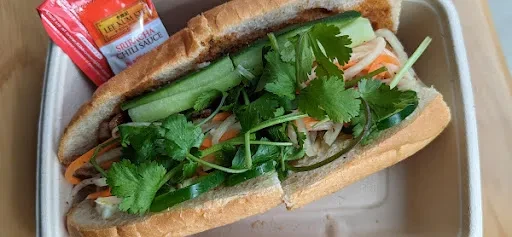 Halved banh mi sandwich in takeout container with Sriracha packet.