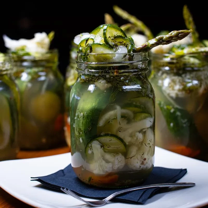 Clear jar of pickled cucumbers, cauliflower, and dill on a table.