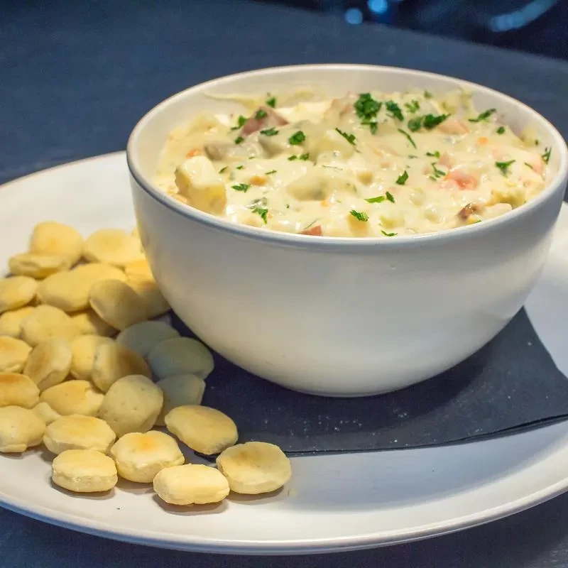Close-up of creamy chowder with potatoes, carrots, ham, parsley, and oyster crackers.