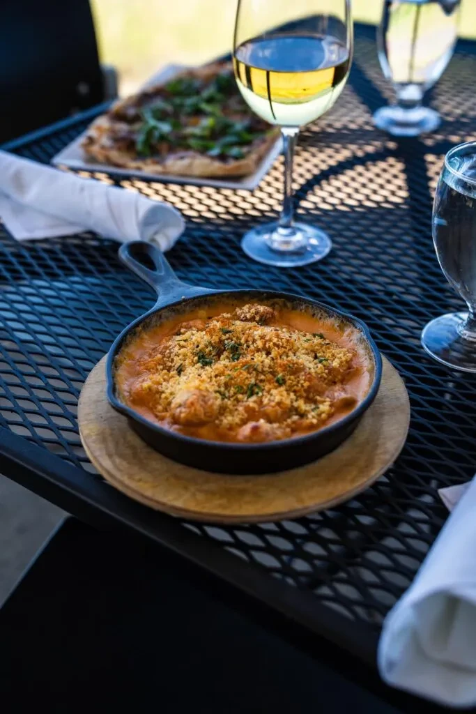 Close-up of baked dish, wine, water on black mesh outdoor table.