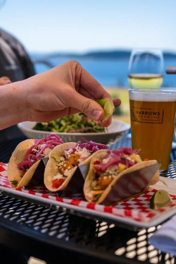 Three tacos with lime being squeezed, outdoors by water.
