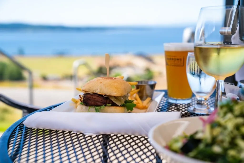 Gourmet burger, fries, wine, beer on outdoor table with water view.