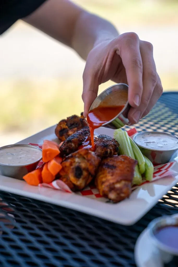 Hand pours red sauce on buffalo wings with celery, carrots, and dip.
