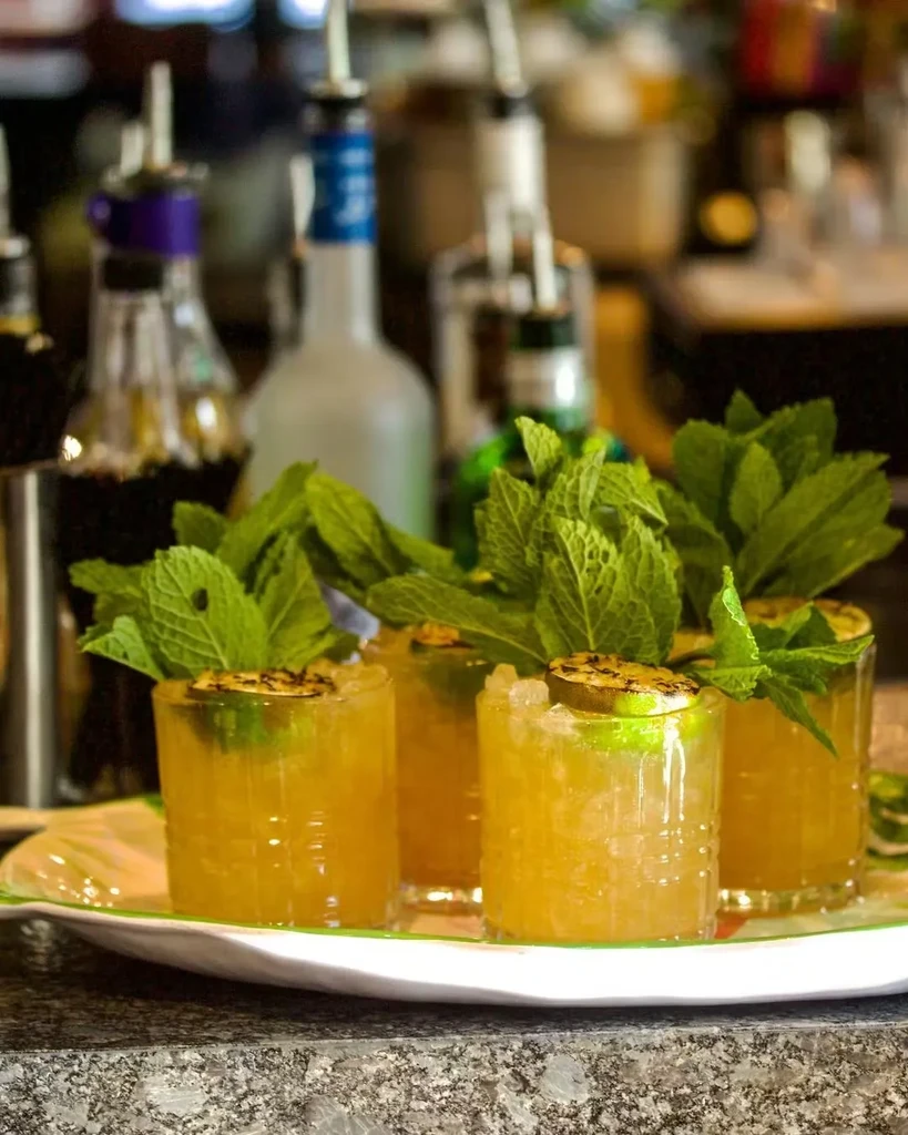Four golden cocktails with mint and charred lime on a bar counter.