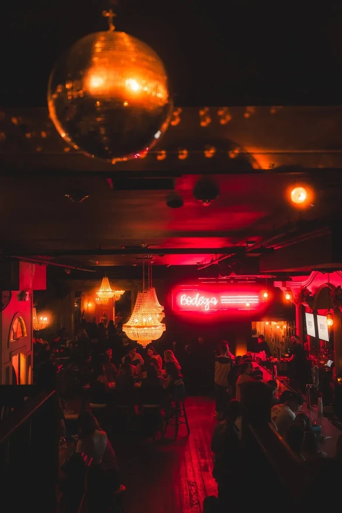 Interior of crowded red-lit bar with 'Bodega' neon and disco ball.