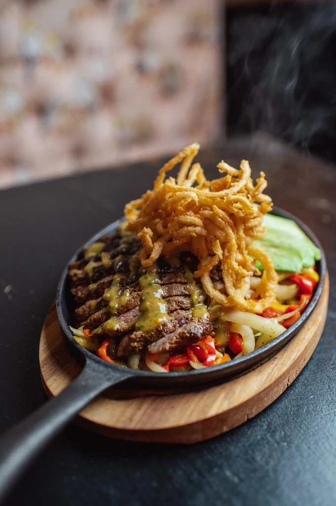 Sizzling meat with fried onions and vegetables in a cast-iron skillet.