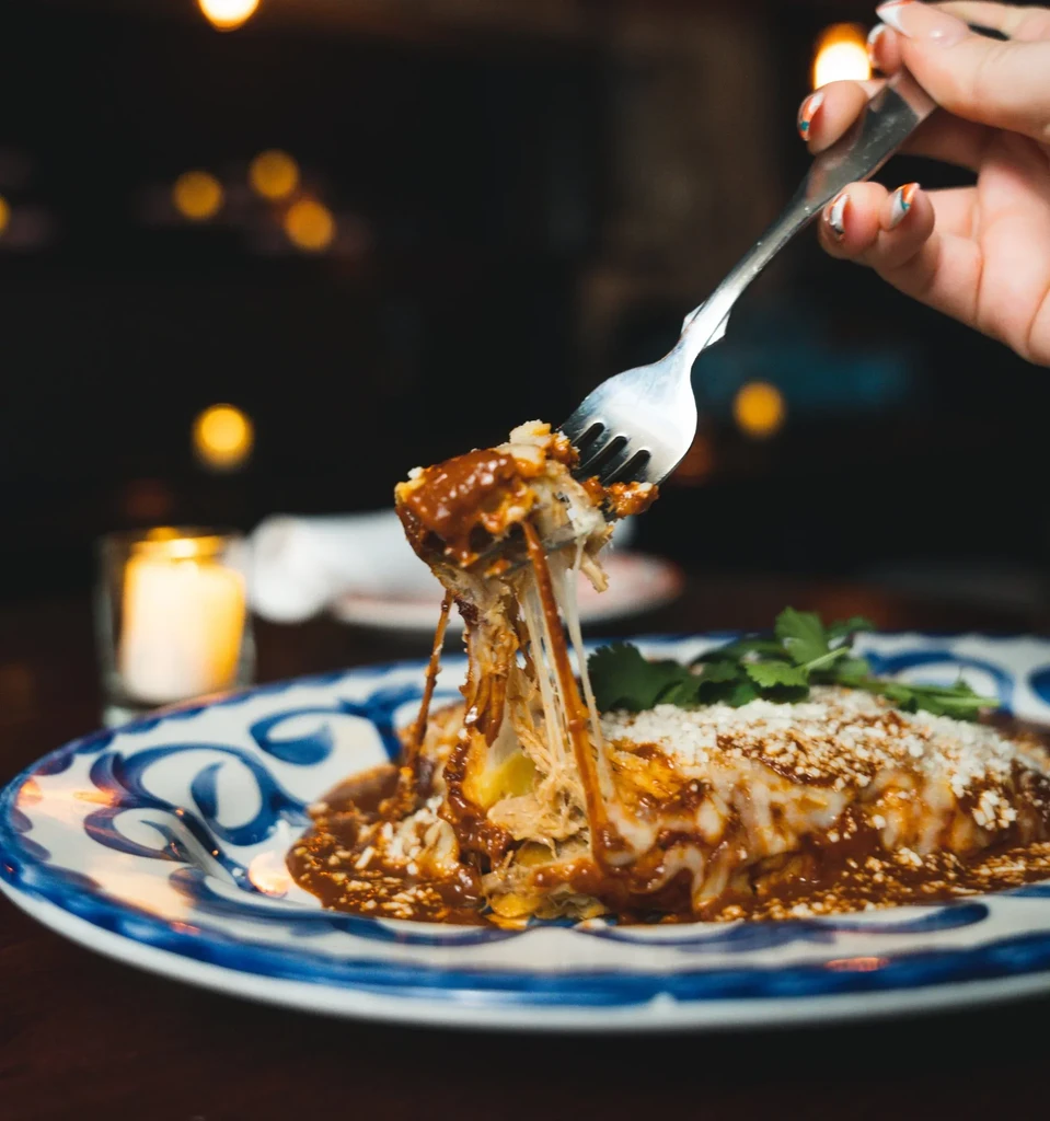 Fork lifts cheesy enchilada from a plate in a cozy restaurant.