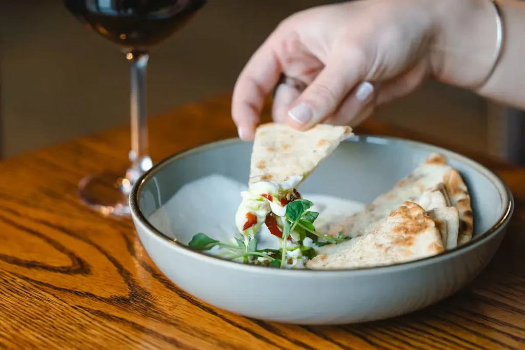 Hand dips flatbread into creamy herbed dip; red wine visible.