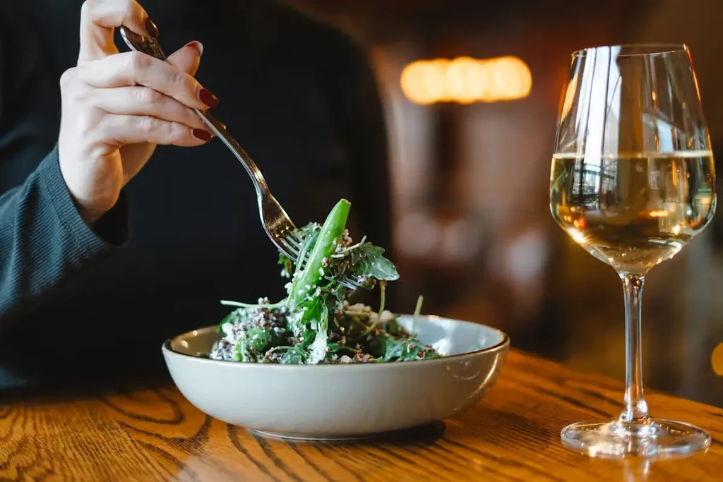 Close-up of hand forking green salad; white wine glass on table.