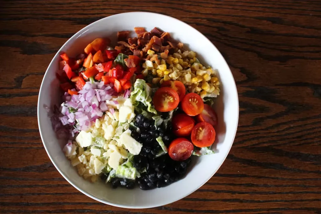 Colorful salad in distinct sections: peppers, corn, tomatoes, cheese, bacon.
