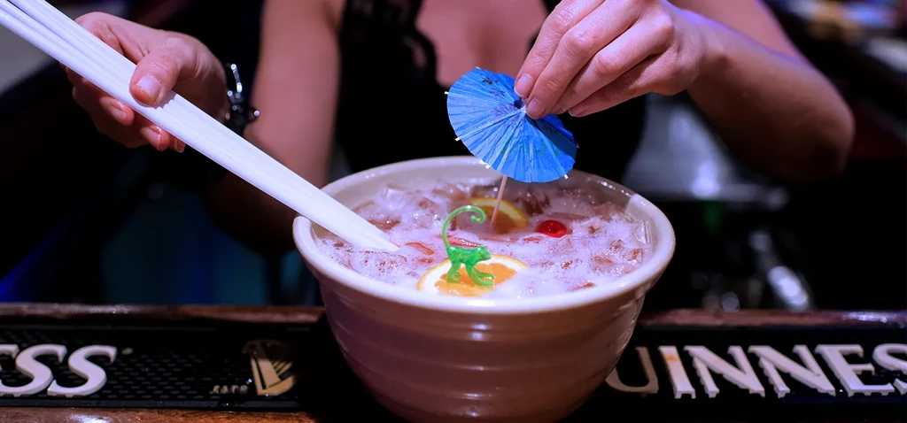Hands adding straws, umbrella to pink iced communal drink with dinosaur.