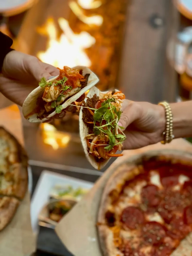 Hands hold colorful meat and vegetable tacos near blurred pizza and flames.