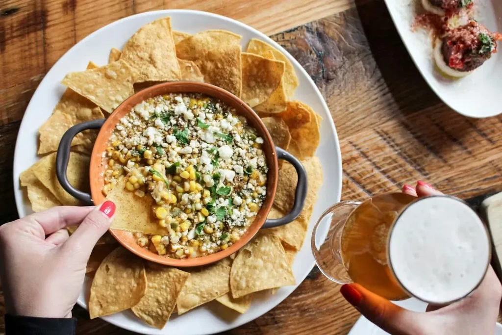 Hand dipping chip into corn dip on rustic table with beer.