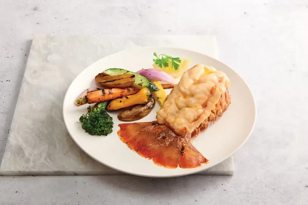 Baked lobster tail with melted topping, colorful roasted vegetables, and lemon.