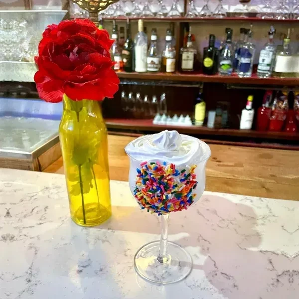 Frothy white drink with whipped cream, sprinkles, and red flower in a bar.