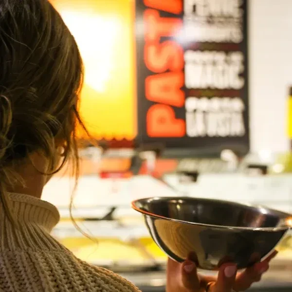 Person's back holds empty steel bowl, blurred red "PASTA" sign.