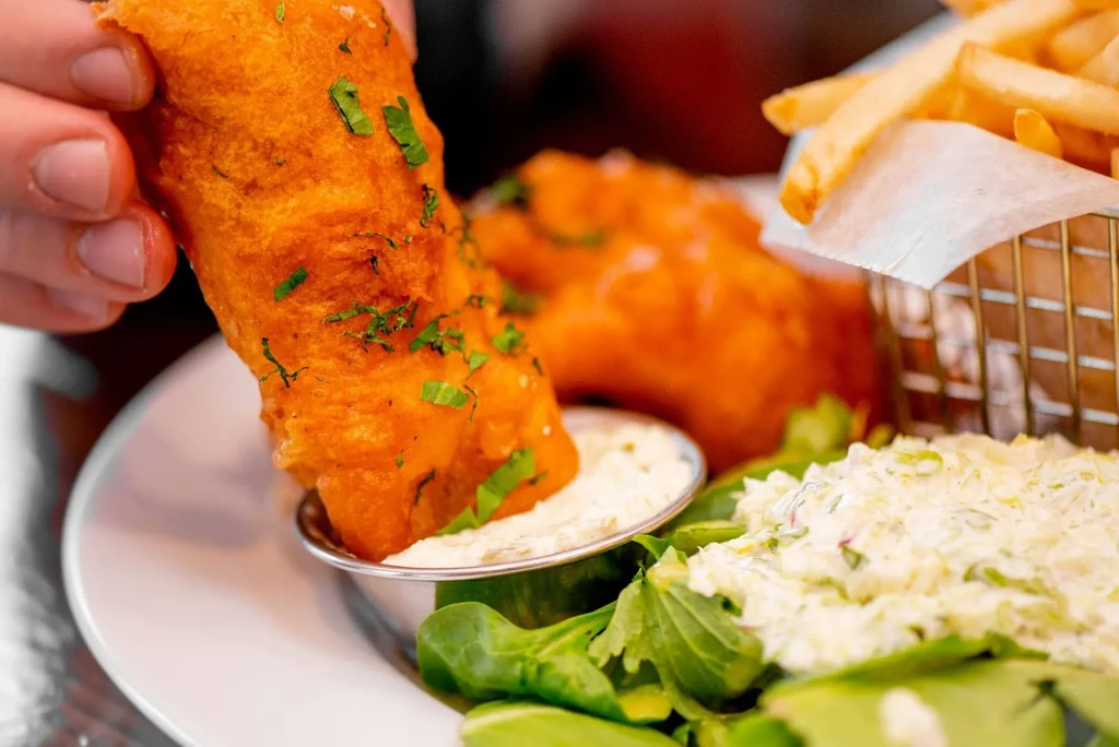 Hand dips herb-speckled fish into tartar sauce.