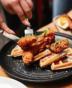 Hands cut into crispy fried chicken and waffles on a black plate.