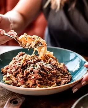 Hands lift cheesy Bolognese pasta with a fork from a blue bowl.