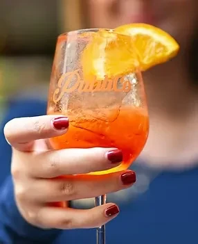 Red-nailed hand holds effervescent orange cocktail with ice and orange.