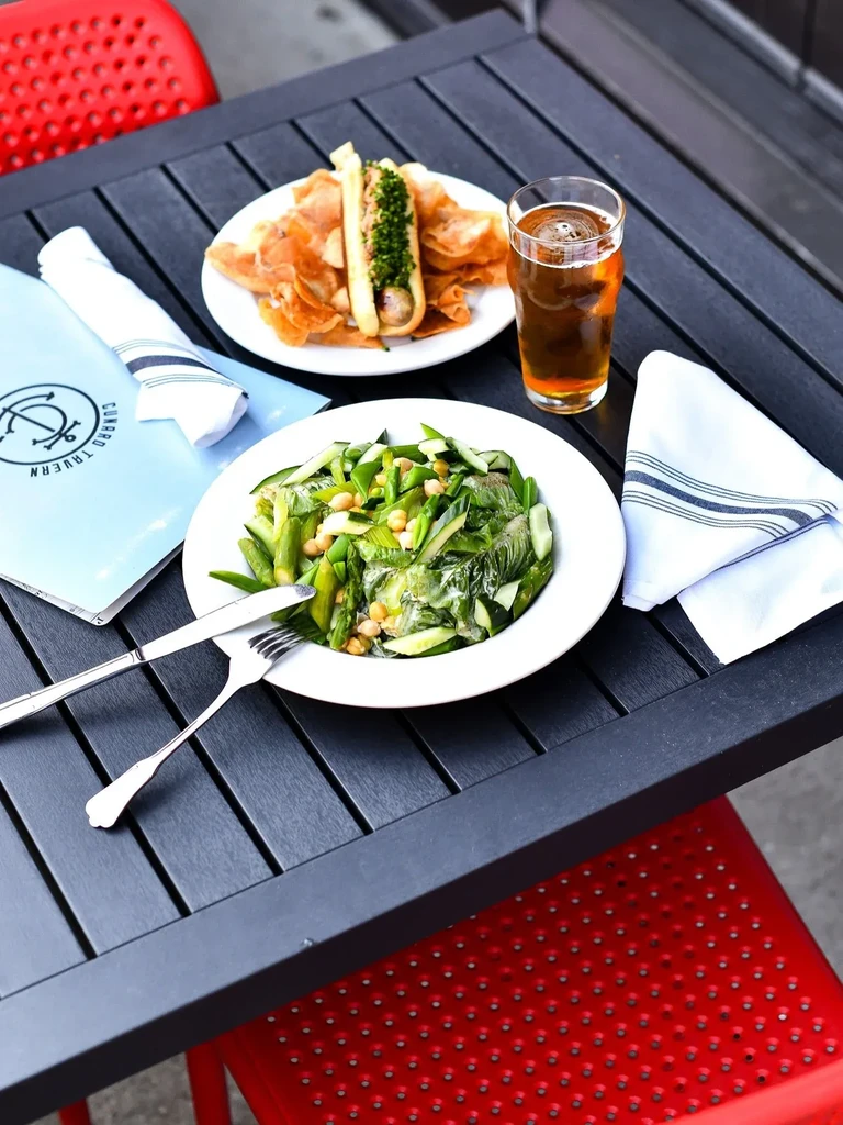 Outdoor dining with a hot dog, salad, beer, and Cumbra Tavern menu.