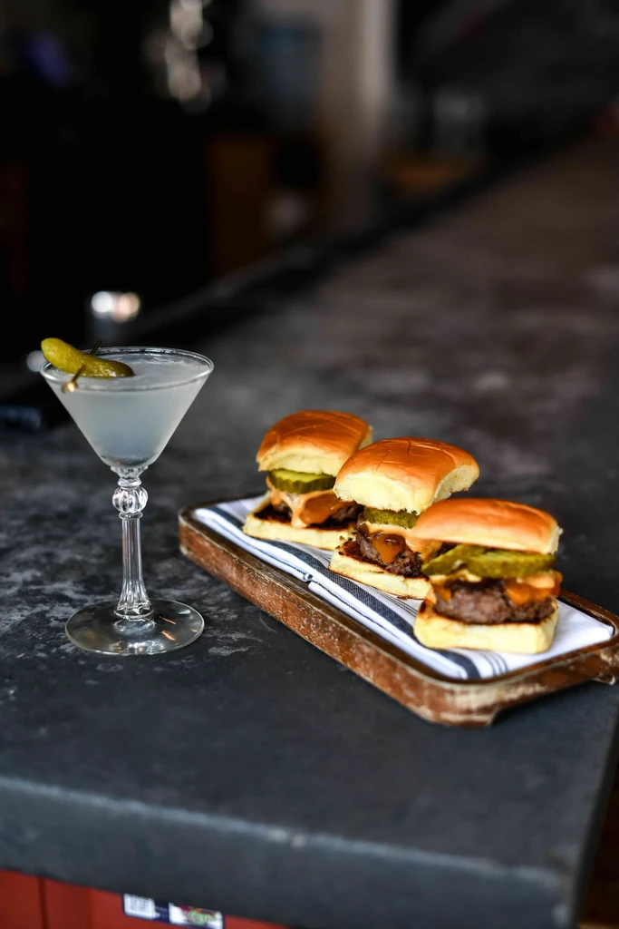Cocktail with gherkin and three mini cheeseburger sliders on a bar.