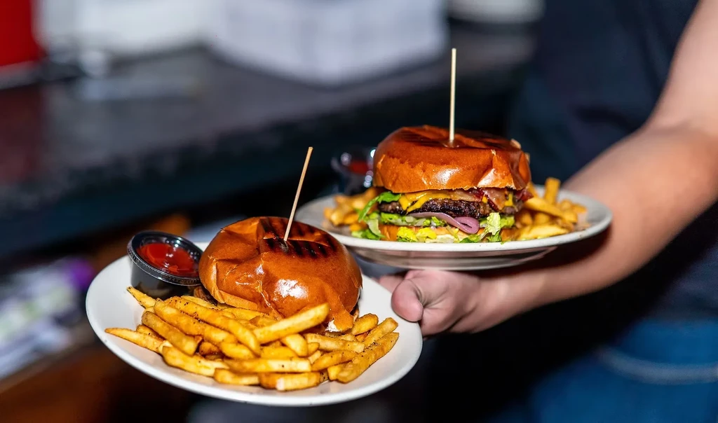Two burgers with fries on plates held by hands.