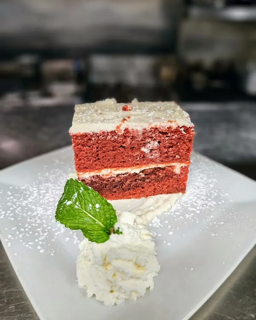 Red velvet cake slice topped with powdered sugar, whipped cream, and mint.