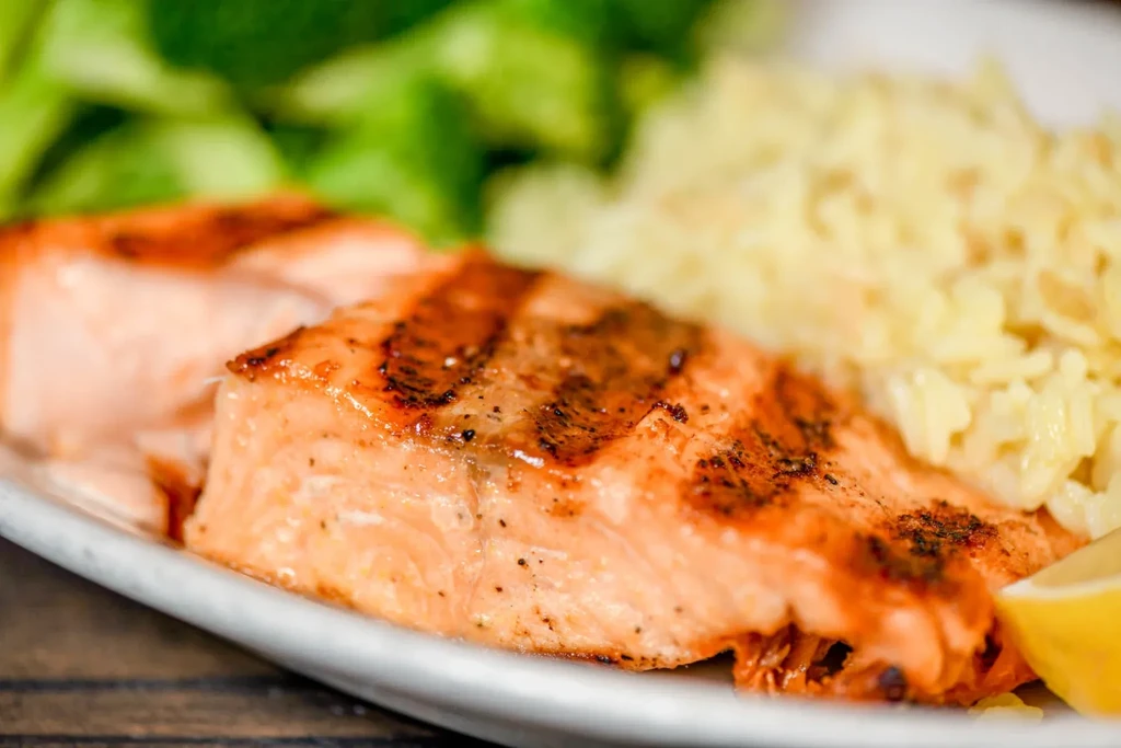 Grilled salmon, yellow rice, lemon, and greens served on a white plate.