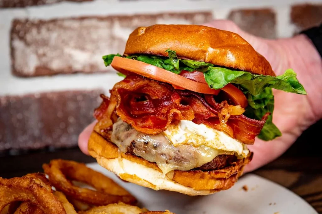Hand holds a brioche burger with beef, egg, bacon, and onion rings.