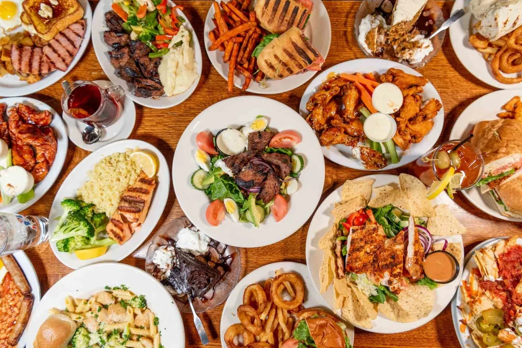 Flat-lay of many diverse restaurant dishes, desserts, and drinks on a table.