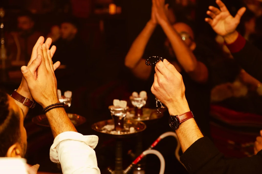 Hands clapping and holding prayer beads at a dimly lit shisha lounge.