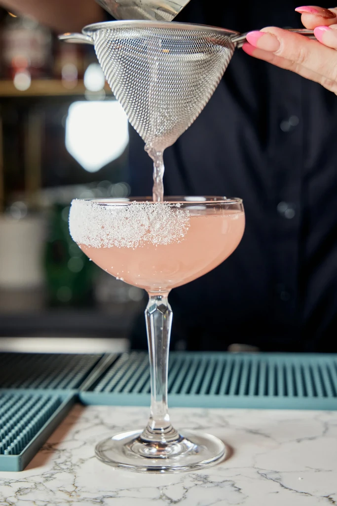 Pink cocktail poured into a sugar-rimmed coupe glass at a bar.