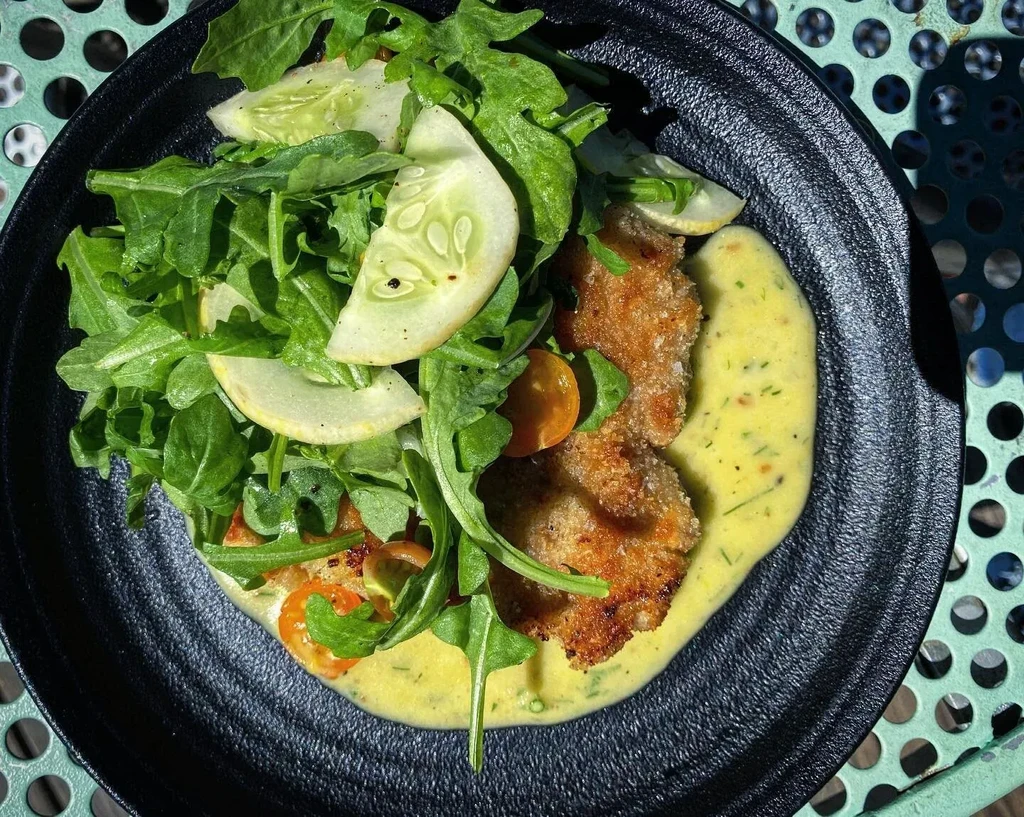 Overhead: Breaded cutlets, green salad, creamy sauce on black plate.