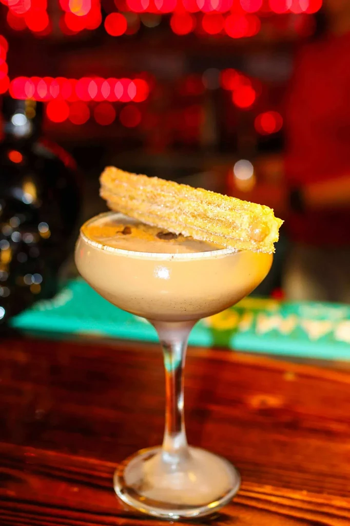 Creamy light brown cocktail in coupe with sugar-dusted churro.