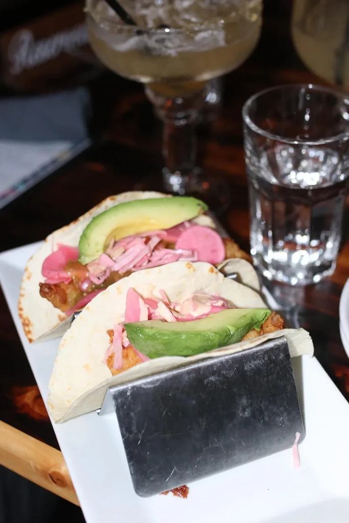 Two vibrantly topped fish tacos on a stand in a dim restaurant.