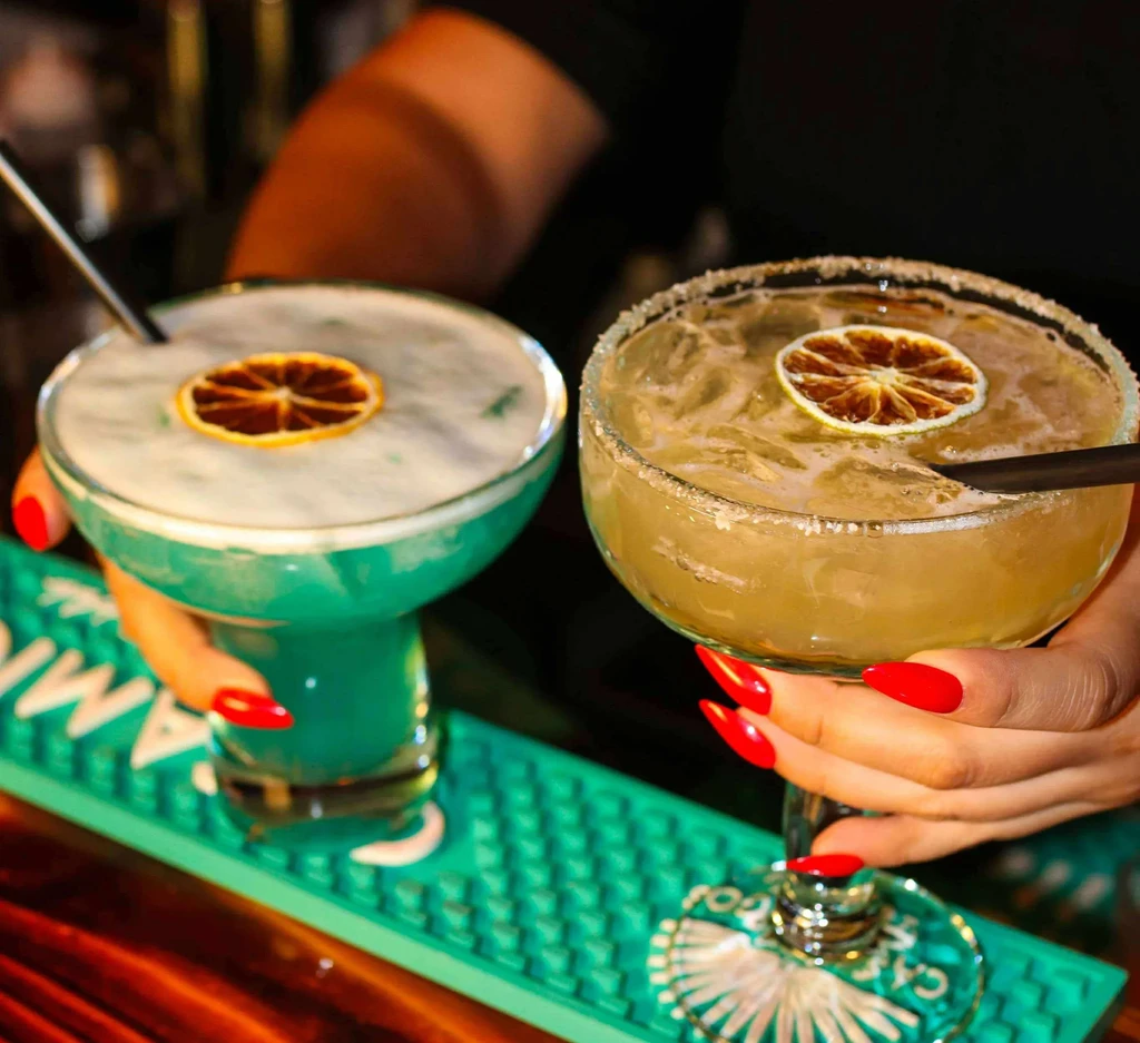 Person holds teal and yellow cocktails with citrus in a bar.