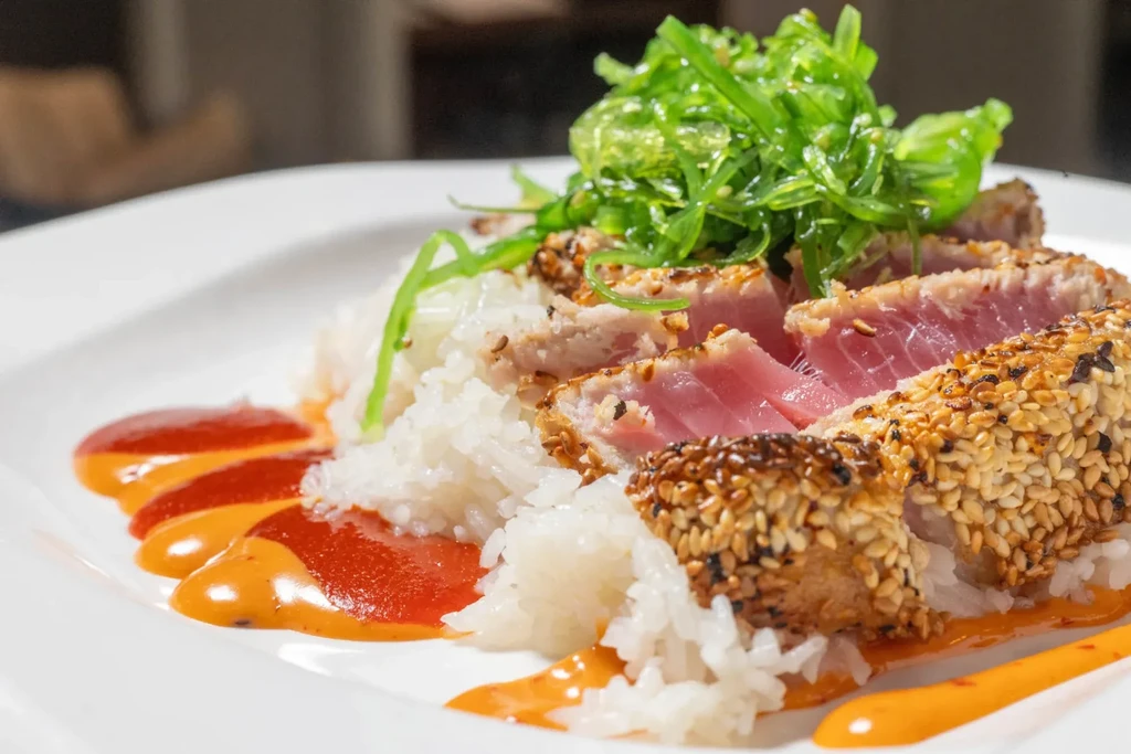 Seared sesame tuna slices on rice with green seaweed salad and sauces.