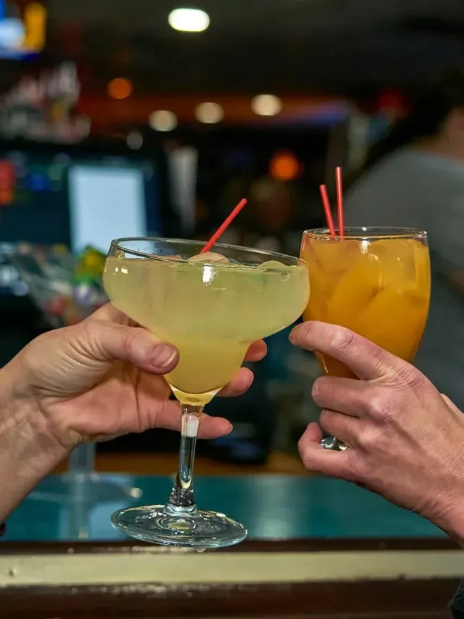 Two hands clink a margarita and an orange drink with straws.