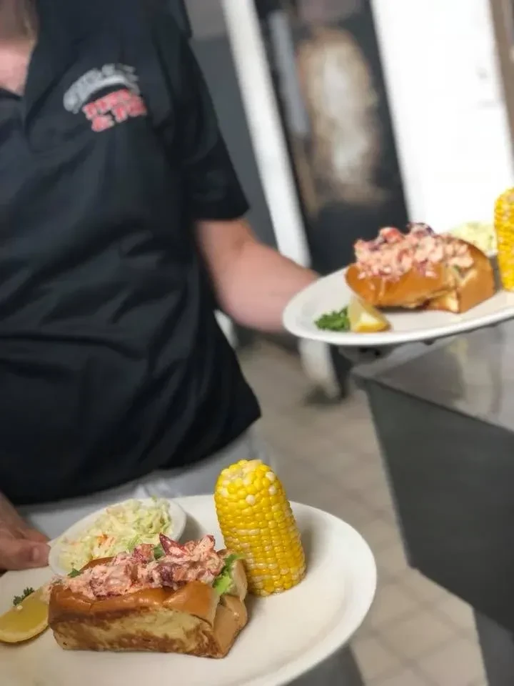 Two plates of lobster rolls with corn and coleslaw, being served.