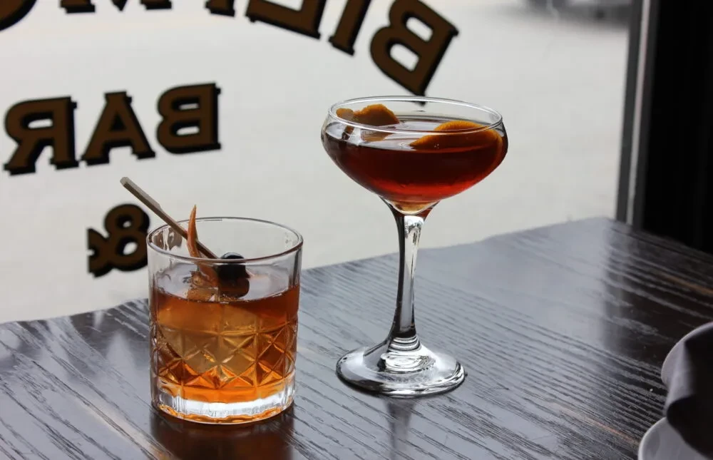 Two garnished cocktails on a dark wooden table, blurred BAR sign.
