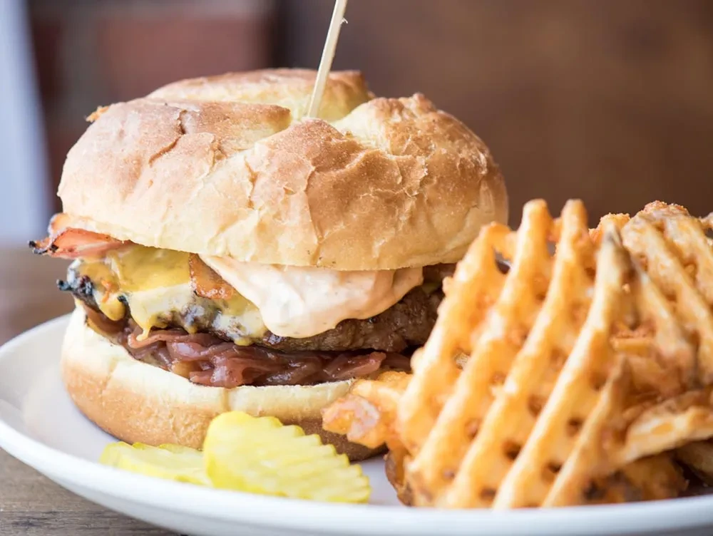 Close-up of a burger, waffle fries, and pickles on a white plate.