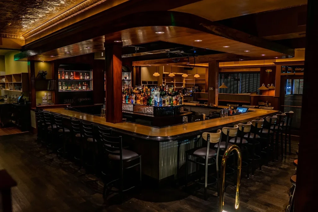 Empty U-shaped wooden bar, brightly lit liquor bottles, ornate ceiling.