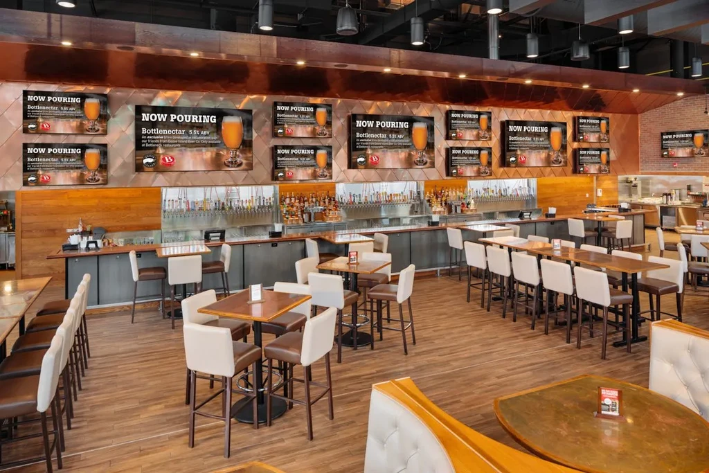 Spacious modern bar interior featuring "NOW POURING" digital display screens.