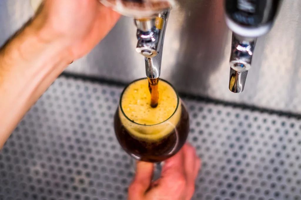 Hands pour dark beverage from a tap into a stemmed glass.