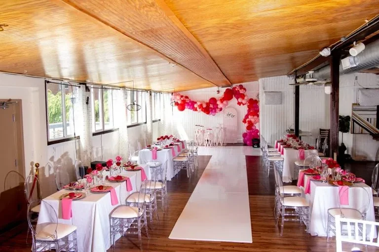 Event room with elegant pink floral tables, ghost chairs, and balloon arch.