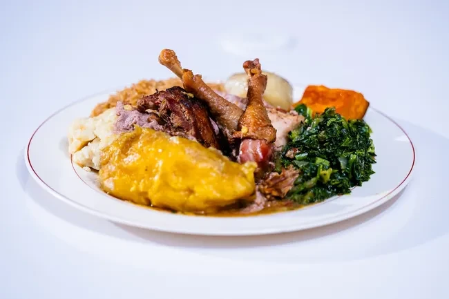 White plate piled high with roasted chicken, fufu, potatoes, and colorful vegetables.